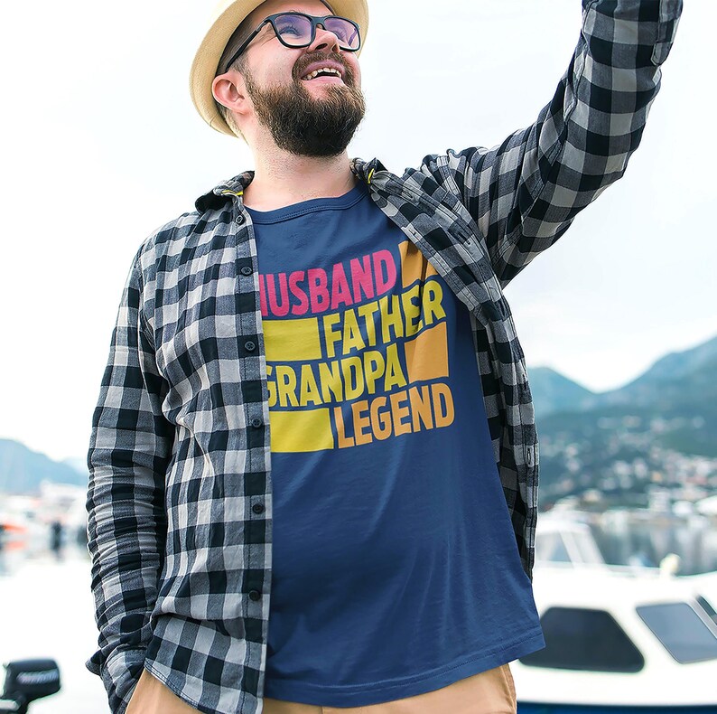 May include: Navy blue t-shirt with the words "HUSBAND FATHER GRANDPA LEGEND" in a colorful, block-letter design. The person in the image is wearing a plaid shirt and a hat.