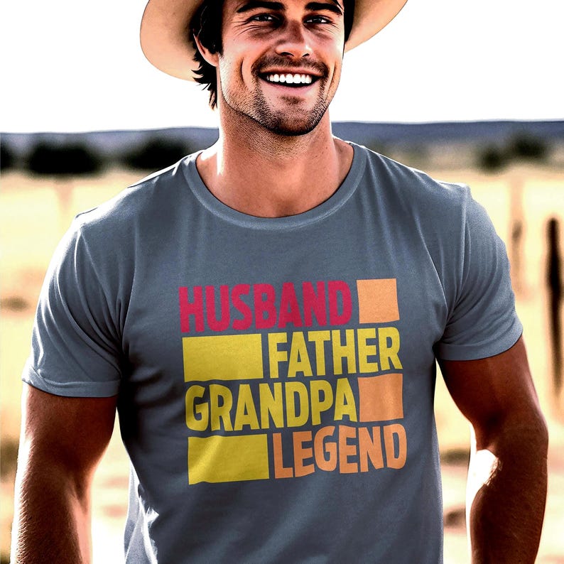 May include: A gray t-shirt with the words "HUSBAND FATHER GRANDPA LEGEND" in a colorful, block-letter design. The shirt is worn by a smiling person wearing a hat, with a blurred outdoor background.