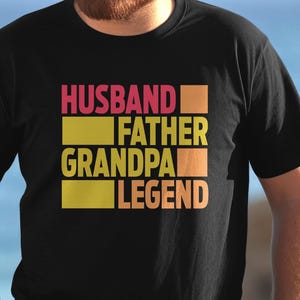 May include: Black t-shirt with the words "HUSBAND FATHER GRANDPA LEGEND" in a stacked design. The text is in a mix of pink, yellow, and orange colors. The shirt is made of a soft material.