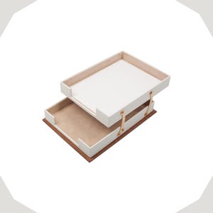 Op de afbeelding: Een bureau-organizer met twee niveaus, voorzien van een witte bovenste lade en een onderste lade met een lichtbruine binnenkant. De lades zijn verbonden door dunne, lichtgekleurde verticale steunen met goudkleurige metalen klinknagels op een donkere houten basis.