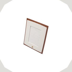 Op de afbeelding: Een rechthoekige fotolijst met een donkerbruine houtnerfrand en een gladde witte passe-partout. De lijst is verticaal geori&euml;nteerd en bevat een kleine, ronde, goudkleurige metalen knop in het midden aan de onderkant van de witte mat.