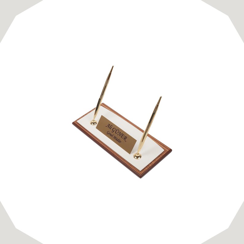 Op de afbeelding: Een rechthoekige bureau-naamplaatset met een houtnerfrand, een wit middenpaneel en twee goudkleurige pennen in rechtopstaande houders. Een goudkleurig metalen plaatje toont de tekst Ali G&Uuml;NER Genel M&uuml;d&uuml;r.