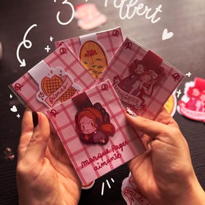 Puede incluir: Una colección de marcapáginas decorativos con varios diseños, incluyendo un gofre con la palabra "friends", e ilustraciones de personajes. Los marcapáginas están empaquetados en plástico transparente y presentan un patrón de cuadros rosas y blancos. El texto "marque Pages aimanté" es visible.