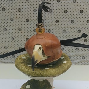May include: A whimsical sculpture featuring a bird skull wearing a brown leather hat, perched atop a green mushroom with white spots. A black cord and decorative accents complete the unique design.