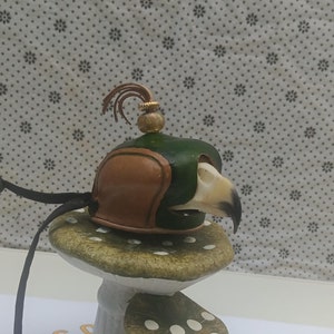 May include: A small, green and brown leather bird skull helmet with a gold top and a brown leather plume. The helmet is sitting on a white mushroom with green and white polka dots.