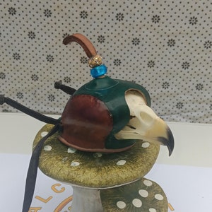May include: A small, brown and green leather and bone bird skull with a blue and gold beaded headpiece, sitting on a green and white ceramic mushroom.