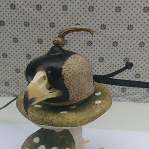 May include: A ceramic mushroom figurine with a bird skull wearing a leather helmet with a feather plume. The mushroom is green with white polka dots.