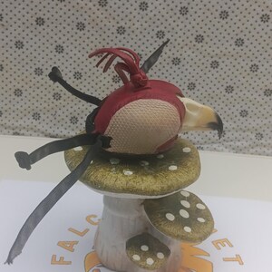 May include: A red and white leather bird figurine perched on a green and white ceramic mushroom. The bird has black leather wings and a long beak.