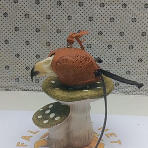 May include: A small, brown leather pouch shaped like a bird skull, with a black cord hanging from it. The pouch is sitting on a green and white mushroom-shaped ceramic stand.