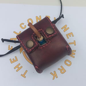 May include: A burgundy leather pouch with a flap closure and a brown leather strap. The pouch has a red stitching detail and two brass snaps. The pouch is hanging from a black cord.