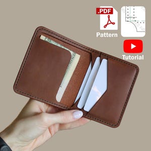 May include: Open brown leather wallet with a cash pocket and card slots. The wallet is held open, revealing a dollar bill and several white cards. Icons for a PDF pattern and a YouTube tutorial are in the background.