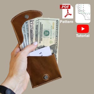 Pode incluir: Uma carteira de couro marrom com fecho magnético, contendo várias denominações de moeda americana e um cartão branco. A carteira está aberta, mostrando o conteúdo. A imagem também inclui ícones para um padrão PDF e um tutorial.