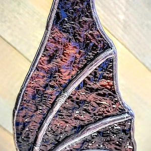 May include: Stained glass bat wing ornament with a textured, multi-colored design. The wing features shades of purple, blue, and orange, framed by a silver-colored metal. The ornament is suspended by a metal loop.