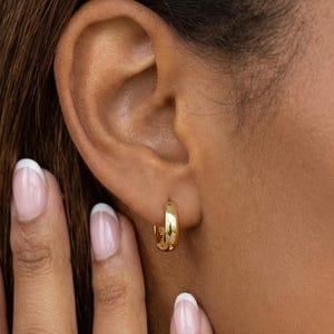 Może przedstawiać: Zbliżenie ucha z małym, złotym kolczykiem w kształcie koła. Kolczyk ma średnicę około 10 mm. Widoczna jest dłoń z zadbanymi paznokciami. Odcień skóry jest średni. Tekst "10mm" jest na zdjęciu.