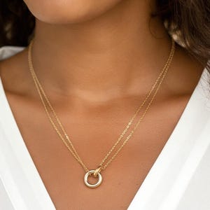 May include: A gold necklace featuring two fine chains and a circular pendant. The pendant has a ring design. The necklace is worn against a white top.