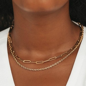 May include: A close-up of a person wearing two gold necklaces. The top necklace is a chain link style, and the bottom necklace is a rope chain style.