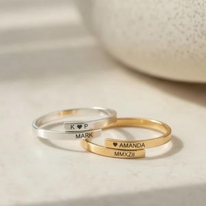 May include: Two personalized wrap rings, one silver and one gold. The silver ring is engraved with "K ♥ P" and "MARK". The gold ring is engraved with "♥ AMANDA" and "MMXXII". The rings are displayed on a white surface.
