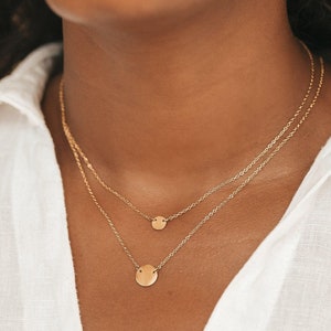 Op de afbeelding: Twee gouden kettingen met ronde hangers. De kettingen zijn gelaagd, met de kleinere hanger op de bovenste ketting en de grotere hanger op de onderste ketting.