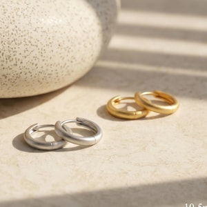 May include: Two pairs of hoop earrings, one pair in silver and the other in gold. The earrings are 10.5mm in diameter and have a simple, minimalist design. The earrings are displayed on a light-colored surface.