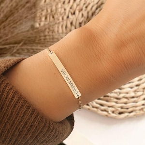 May include: A gold bar bracelet with a delicate chain. The bar has the Roman numeral date "VIII. XII. MMXVI" engraved on it.