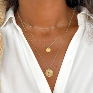 May include: A layered gold necklace with two circular pendants. The larger pendant has a textured surface, while the smaller pendant is smooth.