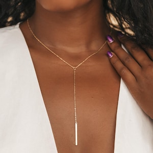 Peut inclure: Un collier en forme de Y de couleur or avec un pendentif barre verticale. Le collier est délicat et tombe bas, créant un look simple mais élégant. Le modèle porte un haut blanc.