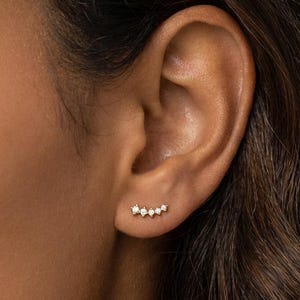 May include: A close-up of an ear with a curved ear climber earring. The earring has a series of small, sparkling gemstones, forming a subtle, elegant arc. The jewelry is set against the backdrop of the person's skin. The gemstones are likely to be set in a gold or silver setting.