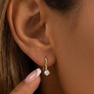 May include: A close-up of a gold hoop earring with a small, square-cut diamond. The earring is worn on the ear. The person's hand is visible, with a French manicure. The earring is a simple, elegant design.