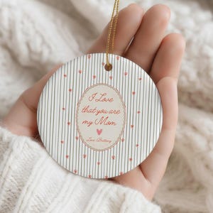 May include: A round ceramic ornament with a gold string for hanging. The ornament has a striped background with small red heart accents. The center features the text "I Love that you are my Mom" in a decorative oval.
