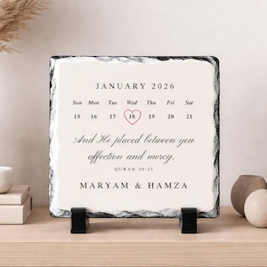 May include: A square slate calendar for January 2026, with a heart marking the 18th. The text reads "And He placed between you affection and mercy. Quran 30:21" and "MARYAM & HAMZA". The calendar is supported by two black stands.