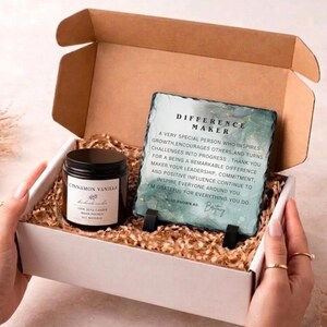 May include: A gift box containing a Cinnamon Vanilla candle and a Difference Maker plaque. The plaque has a teal and gold design with text. The candle is in a black jar with a white label.