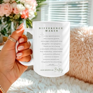 May include: A white ceramic mug with a black handle and the text "But First, Coffee" in a bold, black font. The mug is set against a blurred background. The mug appears to be approximately 10 cm tall.