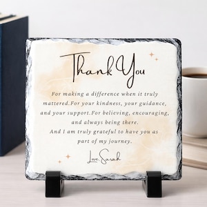 May include: A square slate plaque with the words "Thank You" in elegant script. The plaque features a cream-colored background with a printed message of gratitude and is supported by two black stands. A thoughtful gift.