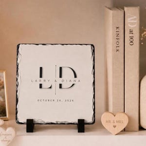May include: A square white stone plaque with rough, jagged edges, displayed on a black stand. The plaque features the black printed text L D, the names Larry & Diana, and the date October 26, 2026. A small wooden heart with the text Mr. & Mrs. sits nearby.