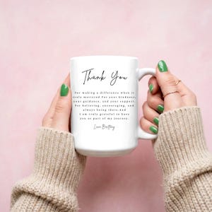 Puede incluir: Una taza de cer&aacute;mica blanca con las palabras "Thank You" en una elegante escritura negra. La taza tambi&eacute;n presenta un mensaje sincero de gratitud. La taza es sostenida por una persona con esmalte de u&ntilde;as verde sobre un fondo rosa suave.