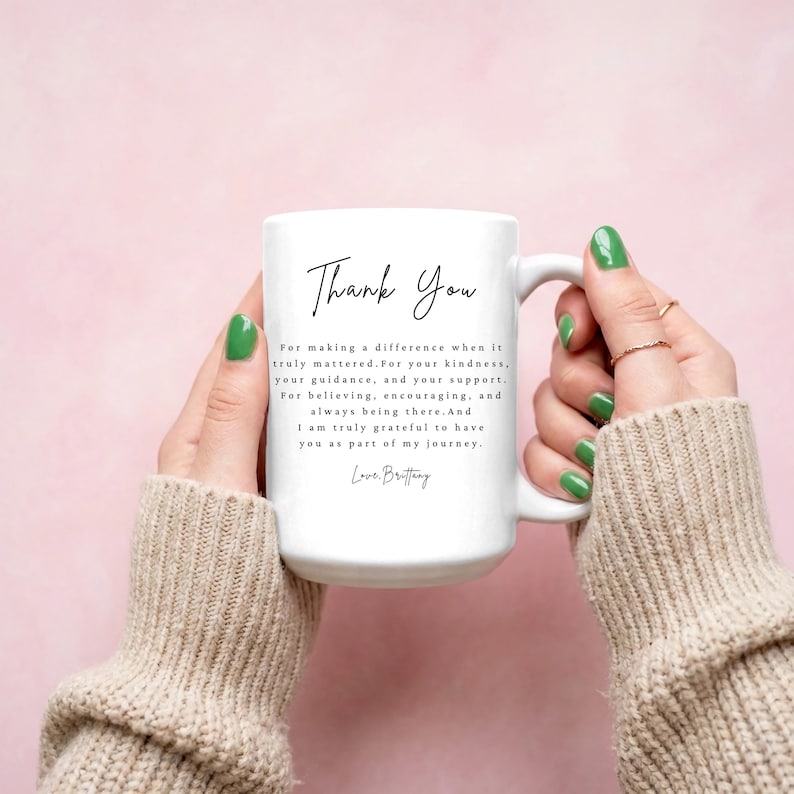 Puede incluir: Una taza de cer&aacute;mica blanca con las palabras "Thank You" en una elegante escritura negra. La taza tambi&eacute;n presenta un mensaje sincero de gratitud. La taza es sostenida por una persona con esmalte de u&ntilde;as verde sobre un fondo rosa suave.