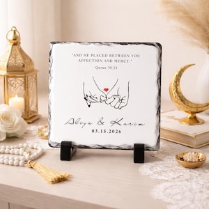 May include: A square, white stone plaque with a black border, featuring the text "AND HE PLACED BETWEEN YOU AFFECTION AND MERCY." and "Quran 30:21." Below is a drawing of two hands holding a red heart, with the names "Aliya & Karim" and the date "03.15.2026."