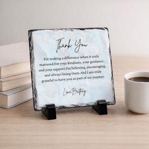 May include: A square slate plaque with the words "Thank You" in elegant script. The plaque features a light blue watercolor-like background and a heartfelt message. It is displayed on a black stand, next to a white mug.