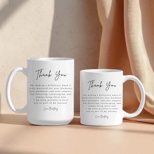 Puede incluir: Dos tazas de cer&aacute;mica blancas con las palabras "Thank You" en escritura elegante. Las tazas tienen un asa curva y un mensaje impreso debajo del texto. El fondo es beige neutro.