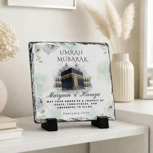 May include: A square slate plaque with the text "UMRAH MUBARAK" and the names "Maryam & Hamza". The image features a depiction of the Kaaba and floral accents. The plaque is supported by two black stands.