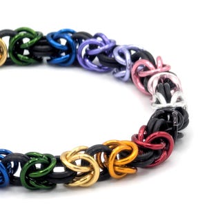 May include: A colorful chainmail bracelet featuring a rainbow of colors. The bracelet is made of interlocking metal rings in shades of green, blue, purple, pink, silver, black, gold, and red. The design is intricate and eye-catching.