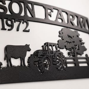 Puede incluir: Un letrero de pared de metal negro con el texto SON FARM y el n&uacute;mero 1972. El dise&ntilde;o incluye una silueta de una vaca, un tractor, un &aacute;rbol y una secci&oacute;n de valla. La pieza de metal recortada se muestra sobre un fondo blanco liso.
