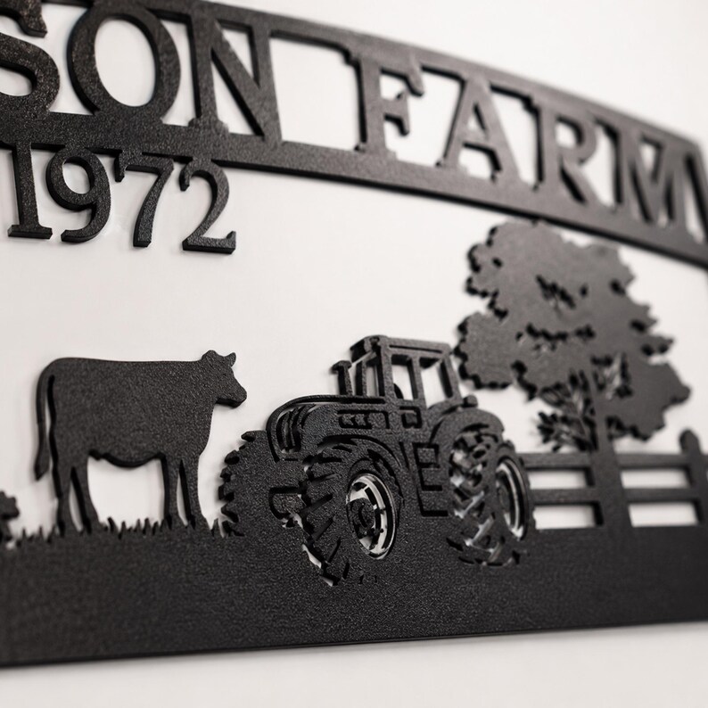 Puede incluir: Un letrero de pared de metal negro con el texto SON FARM y el n&uacute;mero 1972. El dise&ntilde;o incluye una silueta de una vaca, un tractor, un &aacute;rbol y una secci&oacute;n de valla. La pieza de metal recortada se muestra sobre un fondo blanco liso.