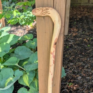 Könnte beinhalten: Ein handgefertigter Holzspazierstock mit gebogenem Griff und natürlichem Finish. Der Stock hat eine hellbraune Farbe mit dunkleren braunen Flecken und eine Gummispitze. Der Griff ist mit einem strukturierten Design geschnitzt.