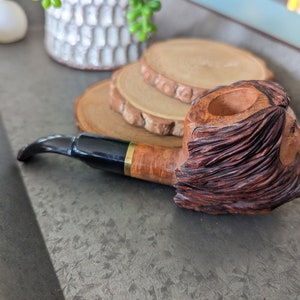 Peut inclure: Une pipe à fumer en bois avec un design sculpté détaillé. La pipe a un embout noir et or et une coupe avec une finition texturée en bois.