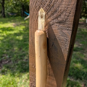 May include: A handcrafted wooden wand with a clear, yellow crystal point. The wand is made of light-coloured wood and is held against a weathered, dark brown wooden post. The background is a blurred view of green grass and trees.