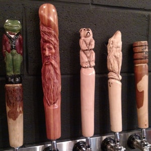 May include: Five wooden beer tap handles with different carvings. The handles are all about 10 inches long and feature a frog, a bearded man, a bear, a wolf, and a stylized design.