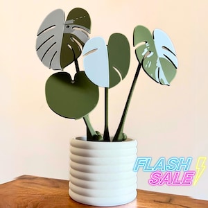 May include: A white ceramic planter with a ribbed design, holding a faux plant with green and white leaves. The leaves are shaped like monstera leaves.