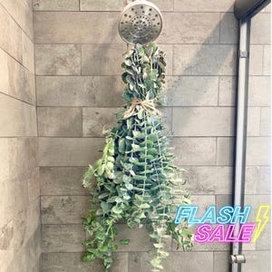 May include: A bunch of eucalyptus leaves hanging from a shower head in a tiled bathroom. The leaves are green and gray and are tied together with twine. The shower head is silver and has a round shape.