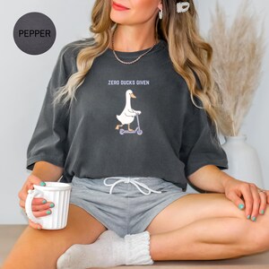 May include: Pepper-colored t-shirt with the text "ZERO DUCKS GIVEN" above a cartoon goose riding a scooter. The shirt is a dark gray color and has a relaxed fit. The goose is white with an orange beak and feet.